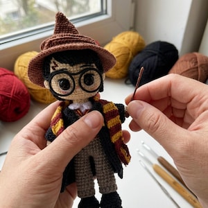 May include: A crocheted doll featuring black hair, round black glasses, and a lightning bolt scar on the forehead. The doll wears a brown pointed hat, a black robe, and a red and yellow striped scarf, while holding a thin brown wand in one hand.