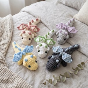 May include: Five small, handmade crochet dragon plushies in various colours including cream with pink wings, light green and white, light purple and grey, yellow with blue wings, and dark grey with light blue wings, each featuring black eyes.