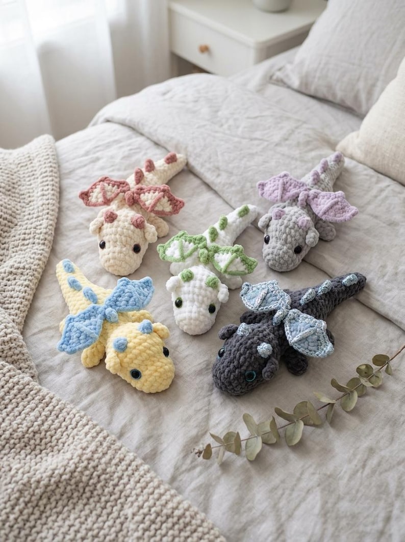 May include: Five small, handmade crochet dragon plushies in various colours including cream with pink wings, light green and white, light purple and grey, yellow with blue wings, and dark grey with light blue wings, each featuring black eyes.