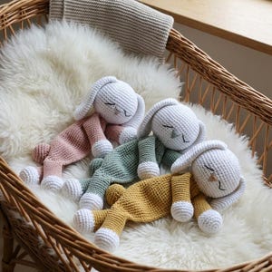 May include: Three crocheted bunny loveys with white heads, long ears, and closed eyes. Each bunny has a solid-colored body in dusty pink, sage green, or mustard yellow, with white hands and feet. The bunnies are lying down in a row.