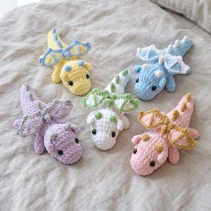 Puede incluir: Cinco pequeños peluches de dragón tejidos a mano en colores pastel. Cada dragón presenta un cuerpo de hilo suave y texturizado, alas pequeñas y ojos de plástico oscuro. La colección incluye un dragón amarillo, azul claro, lavanda, blanco y rosa.