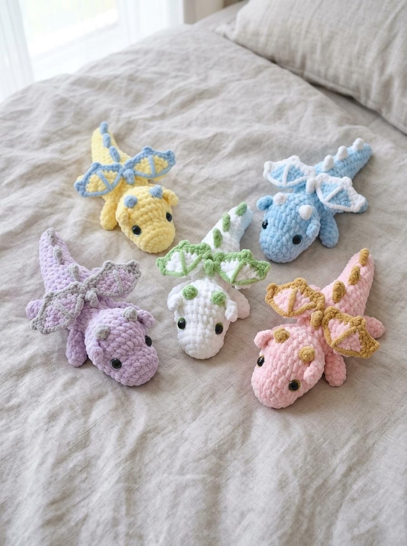 May include: Five small, handmade crochet dragon plushies in pastel colours. Each dragon features a soft, textured yarn body, small wings, and dark plastic eyes. The collection includes a yellow, light blue, lavender, white with green accents, and pink dragon.