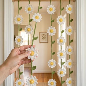 May include: A doorway curtain featuring multiple vertical strands of crocheted white daisy flowers with yellow centers and green stems with leaves, all suspended from a horizontal wooden rod mounted within a white door frame.