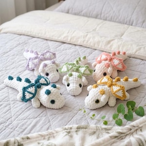 May include: Five small, handmade crochet dragon plushies with white bodies and distinct coloured wings and spikes in lavender, sage green, peach, teal, and mustard yellow. Each soft toy features dark safety eyes and a textured, yarn-based construction.