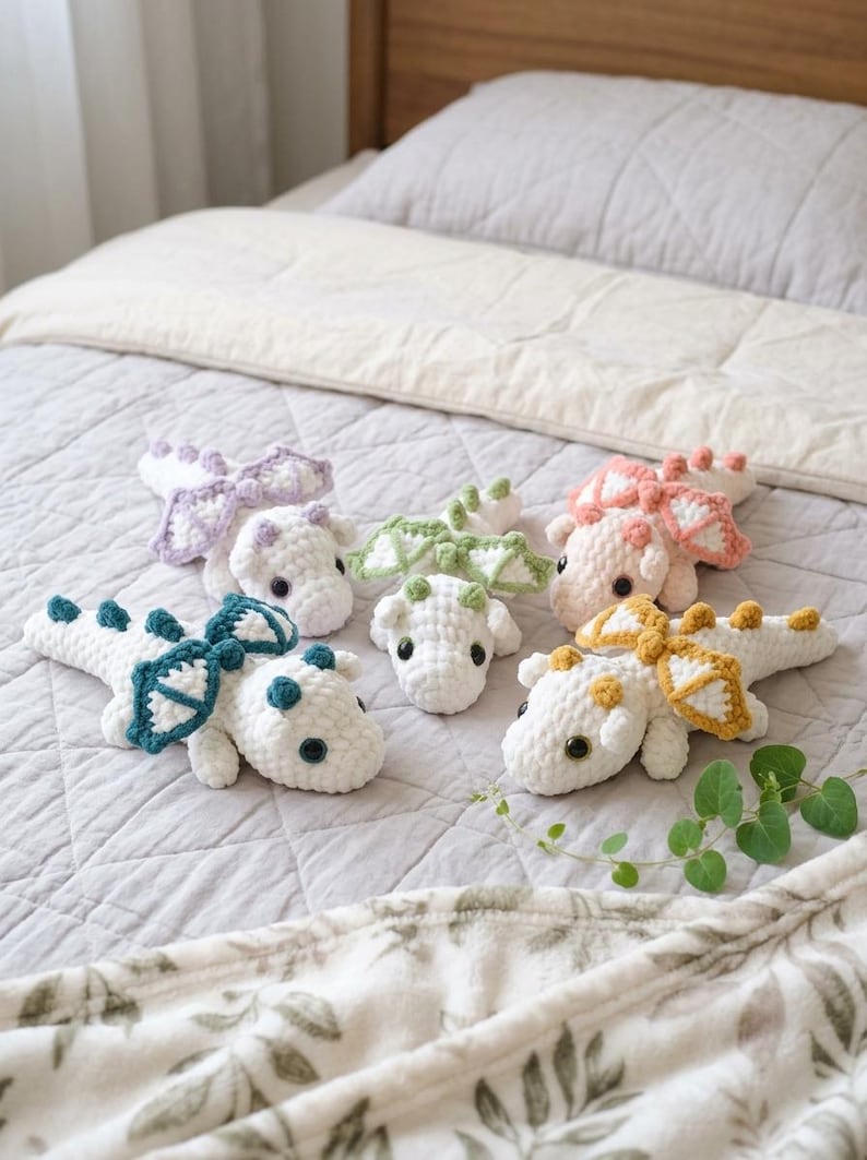 May include: Five small, handmade crochet dragon plushies with white bodies and distinct coloured wings and spikes in lavender, sage green, peach, teal, and mustard yellow. Each soft toy features dark safety eyes and a textured, yarn-based construction.