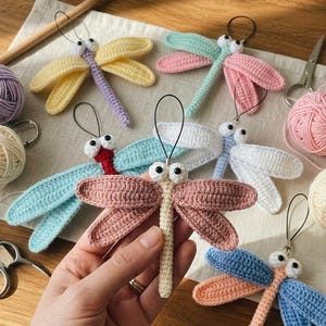 May include: A collection of six handmade crochet dragonfly ornaments, each featuring two pairs of wings, a long segmented body, and two white googly eyes with black pupils. Each dragonfly includes a black cord loop for hanging, crafted in various pastel colors including yellow, pink, blue, and white.