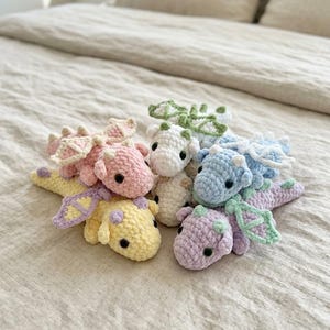 May include: A collection of six small, crocheted dragon plush toys in pastel colours including pink, yellow, white, blue, and purple. Each dragon features soft yarn texture, small wings, rounded bodies, and dark plastic eyes, arranged together in a cluster.