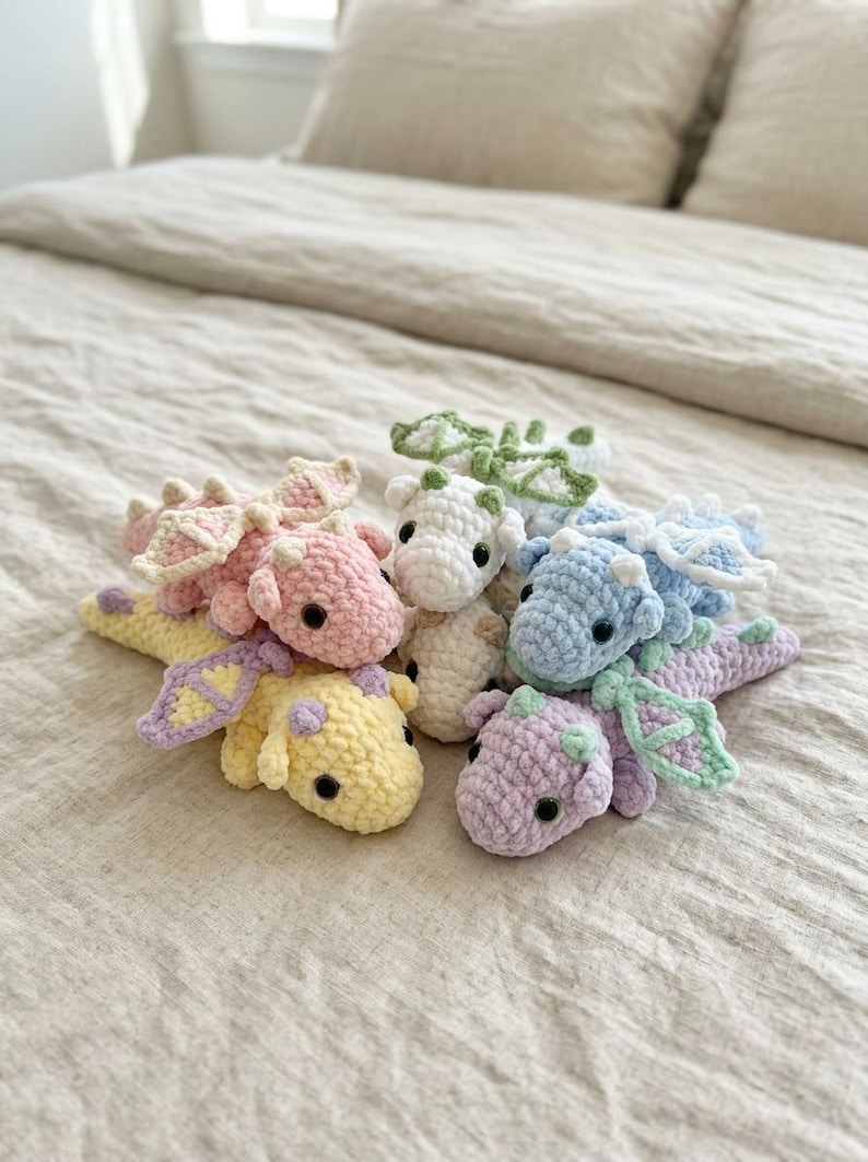 May include: A collection of six small, crocheted dragon plush toys in pastel colours including pink, yellow, white, blue, and purple. Each dragon features soft yarn texture, small wings, rounded bodies, and dark plastic eyes, arranged together in a cluster.