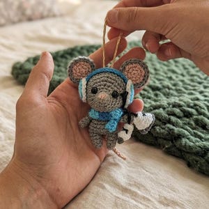 May include: A handmade crochet mouse ornament. The gray mouse wears a blue scarf and earmuffs, with pink accents on the ears. It holds a tiny pair of white and black skis. A gold string is attached for hanging.
