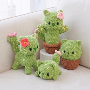 Op de afbeelding: Vier handgemaakte gehaakte knuffels in de vorm van groene katten met cactusachtige texturen, gele steekdetails en kleine roze bloemen. Twee katten staan, één ligt en één zit in een bruine pot, allemaal voorzien van zwarte oogjes.