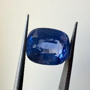 May include: A cushion-cut, deep blue sapphire gemstone held by a pair of tweezers. The sapphire has facets that reflect light, creating a brilliant display of color. The tweezers are dark gray.