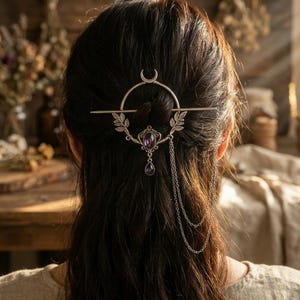 Puede incluir: Accesorio para el cabello plateado con una media luna, una piedra preciosa púrpura ovalada central y cadenas colgantes. La pieza decorativa para el cabello incluye detalles de hojas.