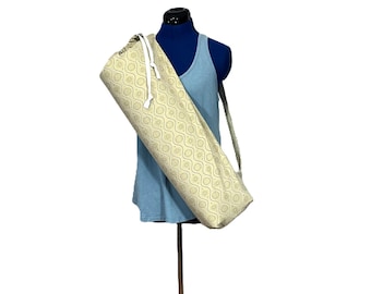 May include: A beige yoga mat bag with a repeating pattern of interconnected circles and lines. The bag has a white drawstring closure and a shoulder strap. The bag is displayed on a blue mannequin wearing a blue tank top.
