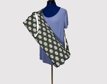 May include: A yoga mat bag with a repeating white floral pattern on a dark green background. The bag has a white drawstring closure and a long strap for carrying. The bag is displayed on a mannequin.