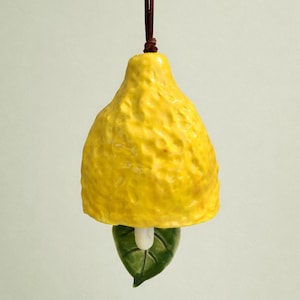 Puede incluir: Adorno de cerámica con forma de limón, con una superficie amarilla texturizada, una hoja verde y un cordón marrón para colgar. El adorno es un artículo decorativo, posiblemente para una cocina o decoración del hogar.