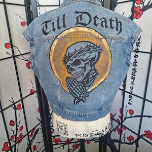 May include: A light blue denim vest featuring a hand-painted design on the back. The artwork depicts a skull wearing a crown of thorns with hands in a praying position, set against a gold circular halo. The text Till Death is printed above.