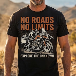 Może przedstawiać: Czarny t-shirt z pomarańczowo-czarną grafiką motocykla i napisem "NO ROADS NO LIMITS EXPLORE THE UNKNOWN". Projekt zawiera łańcuch górski.