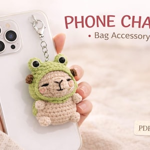 Könnte beinhalten: Ein gehäkelter Telefonanhänger in Form eines Lamas mit grünem Froschhut, an einem Telefon befestigt. Der Anhänger ist beige und grün, mit dem Text "PHONE CHARM - Bag Accessory" im Bild.