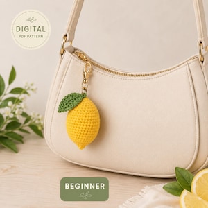 May include: A crocheted yellow lemon keychain with a green leaf attached, hanging from a cream-colored purse. The keychain is a beginner-level digital PDF pattern. The purse has gold-colored hardware and a curved design. The background is a light wood surface.
