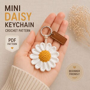 Può includere: Un portachiavi a forma di margherita all'uncinetto con petali bianchi e centro giallo, fissato a un anello in metallo argentato con una piccola etichetta rettangolare in pelle marrone. Il testo MINI DAISY KEYCHAIN CROCHET PATTERN, PDF PATTERN e BEGINNER FRIENDLY è visibile.