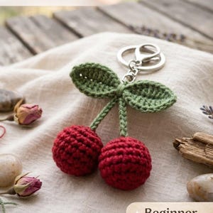 Puede incluir: Un llavero de cereza hecho a mano con dos cerezas rojas, hojas verdes y un llavero plateado. El texto "Crochet Pattern PDF" y "Beginner Friendly" están presentes. El fondo incluye flores secas y una superficie de madera.
