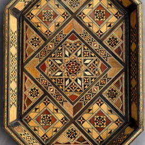 May include: An octagonal wooden tray with an intricate geometric mosaic design. The tray features a dark brown border and a central pattern of diamonds, stars, and squares in shades of brown, red, and beige. The tray is a decorative item.