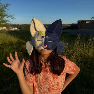 May include: A handmade bunny mask, split into cream and navy blue halves, with large ears. The mask is being worn by a person. The person is wearing a floral print shirt. The background is a grassy field.