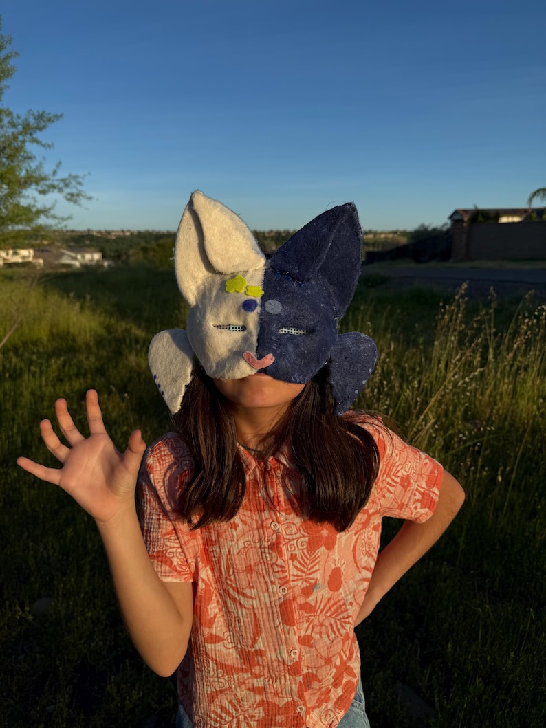May include: A handmade bunny mask, split into cream and navy blue halves, with large ears. The mask is being worn by a person. The person is wearing a floral print shirt. The background is a grassy field.