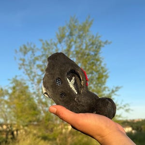May include: A black, plush animal head with white mesh and red detailing. The head has a rounded shape with a snout and ears. The head is held in a hand against a backdrop of green grass and a blue sky.
