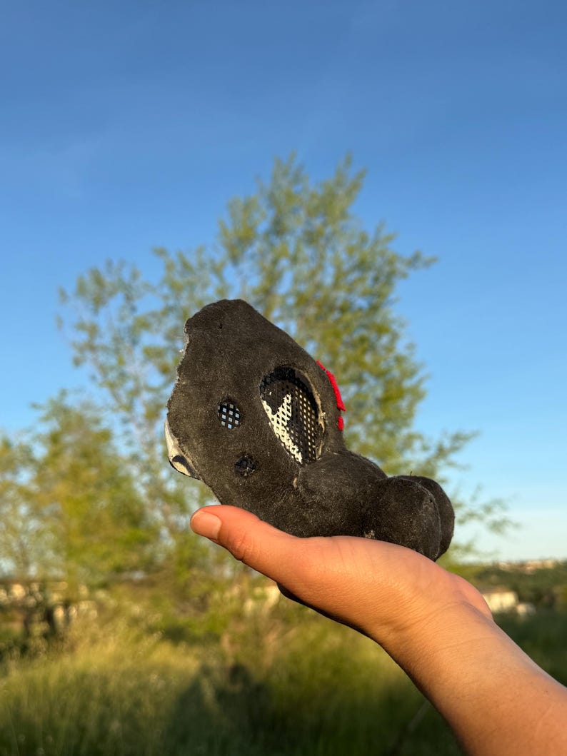 May include: A black, plush animal head with white mesh and red detailing. The head has a rounded shape with a snout and ears. The head is held in a hand against a backdrop of green grass and a blue sky.