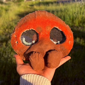 May include: A brown and orange mask with mesh eye coverings and a nose and two lower appendages. The mask is held in a hand against a blurred green background. The mask has a textured, felt-like appearance.