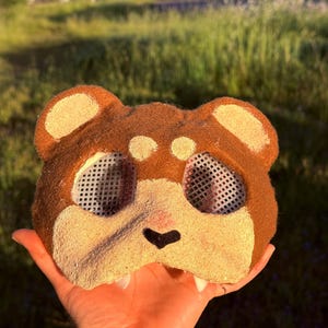 May include: A brown and tan bear mask with mesh eye coverings. The mask has a black nose and tan accents on the ears and cheeks. The mask is held up by a hand.