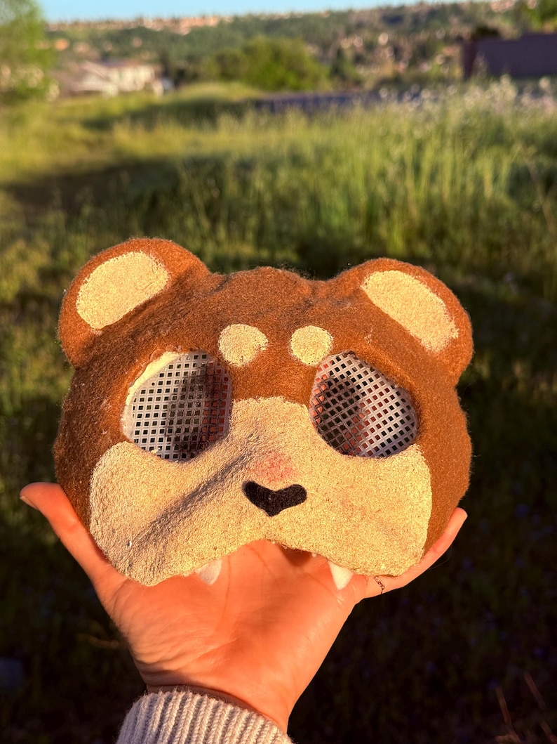 May include: A brown and tan bear mask with mesh eye coverings. The mask has a black nose and tan accents on the ears and cheeks. The mask is held up by a hand.