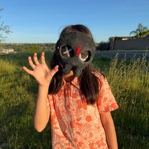 May include: A black mask with white cross and red details. The mask is worn by a person wearing an orange floral print shirt and blue denim shorts. The background is a grassy field and a clear blue sky.