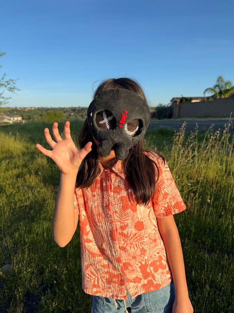 May include: A black mask with white cross and red details. The mask is worn by a person wearing an orange floral print shirt and blue denim shorts. The background is a grassy field and a clear blue sky.