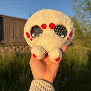 May include: A soft, round, cream-coloured creature with two large, mesh eyes and red markings. The toy has small, rounded feet with red details. The creature is held in a hand against a backdrop of green grass and a building.
