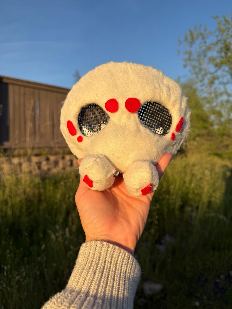 May include: A soft, round, cream-coloured creature with two large, mesh eyes and red markings. The toy has small, rounded feet with red details. The creature is held in a hand against a backdrop of green grass and a building.