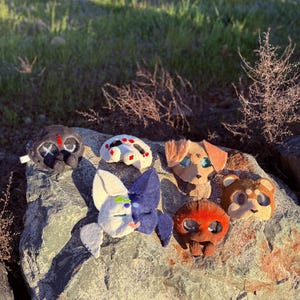 May include: A selection of handmade felt animal masks in various colours and designs, including a spider, dog, bear, and other creatures. The masks are arranged on a grey rock outdoors, with sunlight and greenery in the background.