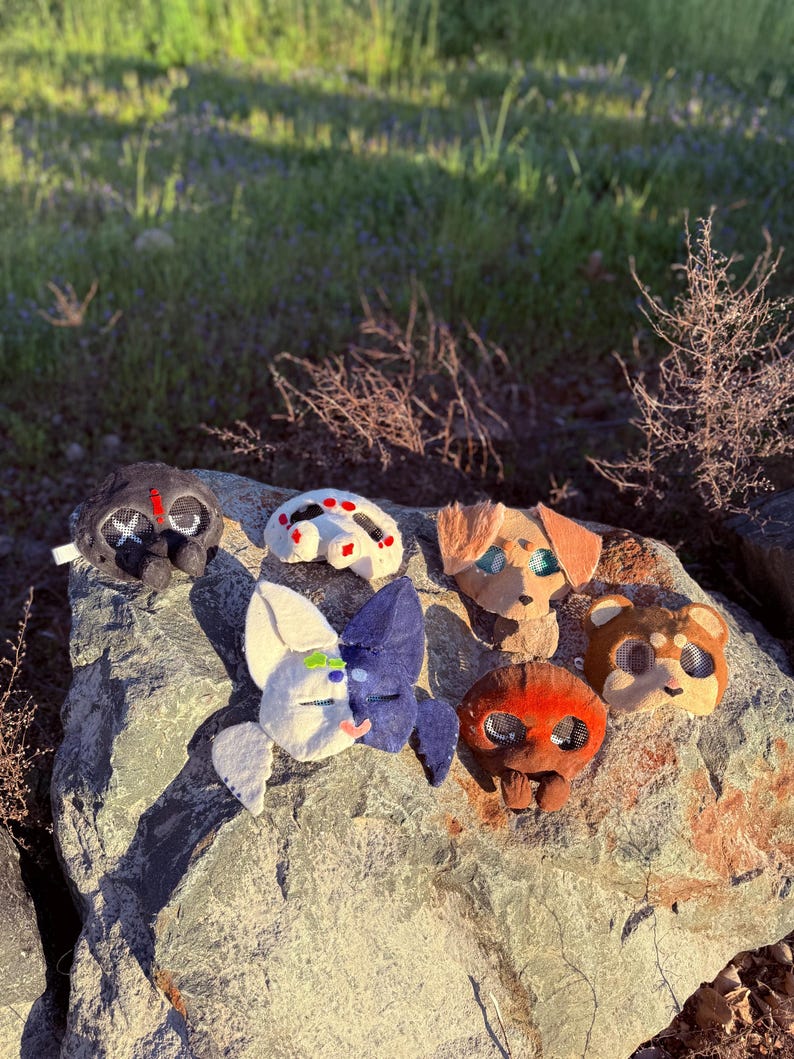 May include: A selection of handmade felt animal masks in various colours and designs, including a spider, dog, bear, and other creatures. The masks are arranged on a grey rock outdoors, with sunlight and greenery in the background.