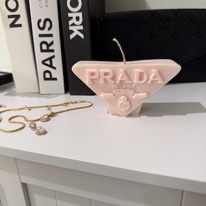 Puede incluir: Una vela rosa pálido con forma de triángulo, con la inscripción "PRADA MILANO DAL 1913". En primer plano, un collar dorado con colgantes con forma de diamante. La vela está sobre una superficie blanca.