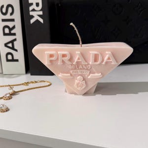 Puede incluir: Una vela rosa pálido en forma de triángulo, con la palabra "PRADA" en relieve. La vela también presenta las palabras "MILANO DAL 1913" y un escudo. Un collar de oro con un colgante de diamantes está en primer plano.