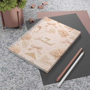 Könnte beinhalten: Ein cremefarbenes Spiralbuch mit floralem Muster und Schleifen. Die Worte "my Journal" sind auf dem Einband aufgedruckt. Zwei Bleistifte, einer rosa und einer weiß, liegen neben dem Notizbuch. Das Notizbuch liegt auf einer grauen Oberfläche.