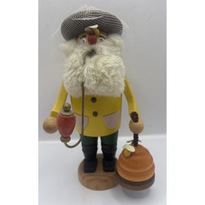 May include: A wooden figurine of a beekeeper, wearing a yellow coat, green pants, and a hat with a veil. The figurine has a white beard and holds a red lantern and a beehive. The base is made of wood.