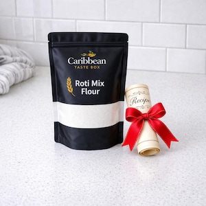 May include: A black bag of "Caribbean Taste Box" Roti Mix Flour with a white stripe. A rolled-up recipe tied with a red bow sits next to the bag. The background is a white tiled wall and a white countertop.