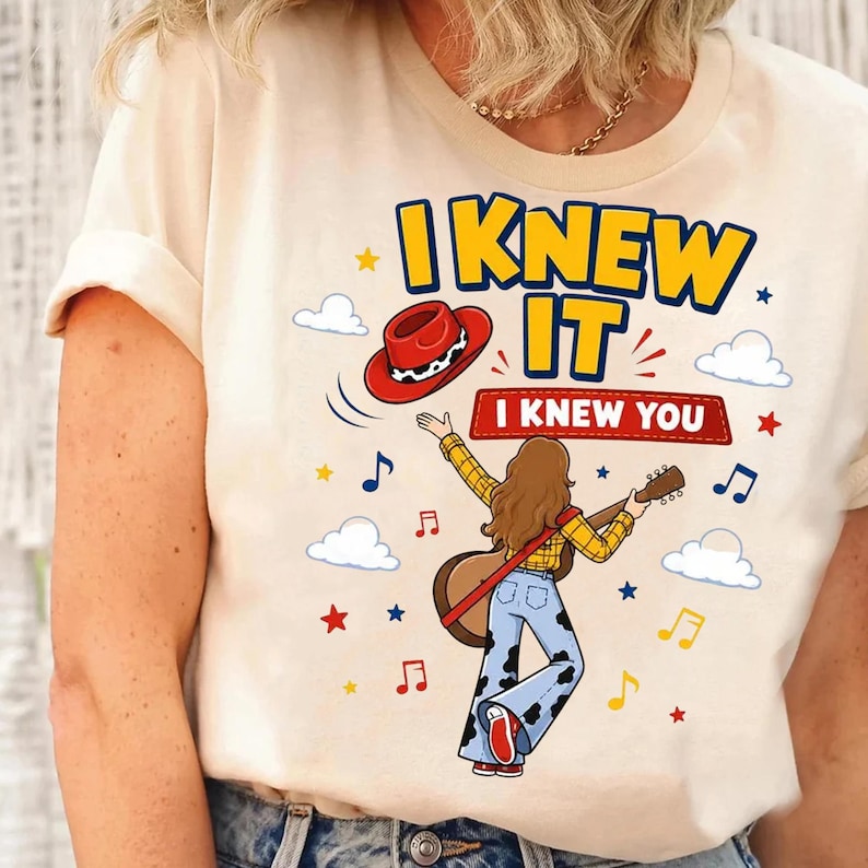 Puede incluir: Una camiseta color crema con un gr&aacute;fico de una persona de pelo largo casta&ntilde;o tocando una guitarra ac&uacute;stica, vistiendo una camisa de cuadros amarilla y pantalones con estampado de vaca. El texto dice I KNEW IT y I KNEW YOU, con sombrero rojo.