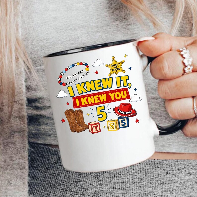 Pu&ograve; includere: Una tazza in ceramica bianca con manico e bordo neri, decorata con una grafica colorata con il testo "I KNEW IT, I KNEW YOU", una stella da sceriffo, un cappello da cowboy, stivali marroni, cubi con "TS 5" e un disegno di braccialetto di perline.