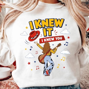 Puede incluir: Una sudadera blanca con un gr&aacute;fico de una persona de pelo largo casta&ntilde;o tocando la guitarra, vistiendo una camisa de cuadros amarillos y pantalones con estampado de vaca. El dise&ntilde;o incluye un sombrero de vaquero rojo, notas musicales, estrellas, nubes y el texto I KNEW IT I KNEW YOU.
