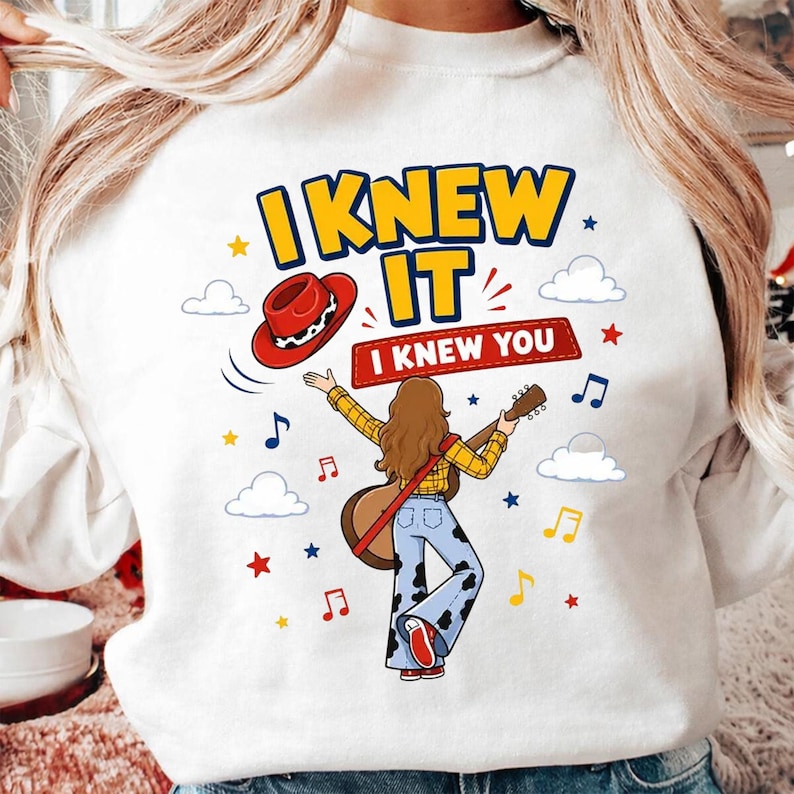 Puede incluir: Una sudadera blanca con un gr&aacute;fico de una persona de pelo largo casta&ntilde;o tocando la guitarra, vistiendo una camisa de cuadros amarillos y pantalones con estampado de vaca. El dise&ntilde;o incluye un sombrero de vaquero rojo, notas musicales, estrellas, nubes y el texto I KNEW IT I KNEW YOU.