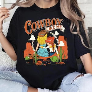 Puede incluir: Una camiseta negra con un estampado gr&aacute;fico que incluye el texto "COWBOY LIKE ME". El dise&ntilde;o muestra a dos figuras con cabello rubio y pelirrojo usando sombreros de vaquero, en un paisaje des&eacute;rtico con formaciones rocosas naranjas y cactus.