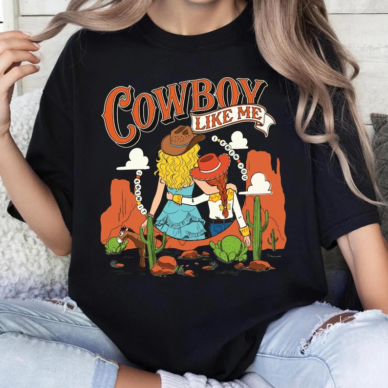 Puede incluir: Una camiseta negra con un estampado gr&aacute;fico que incluye el texto "COWBOY LIKE ME". El dise&ntilde;o muestra a dos figuras con cabello rubio y pelirrojo usando sombreros de vaquero, en un paisaje des&eacute;rtico con formaciones rocosas naranjas y cactus.